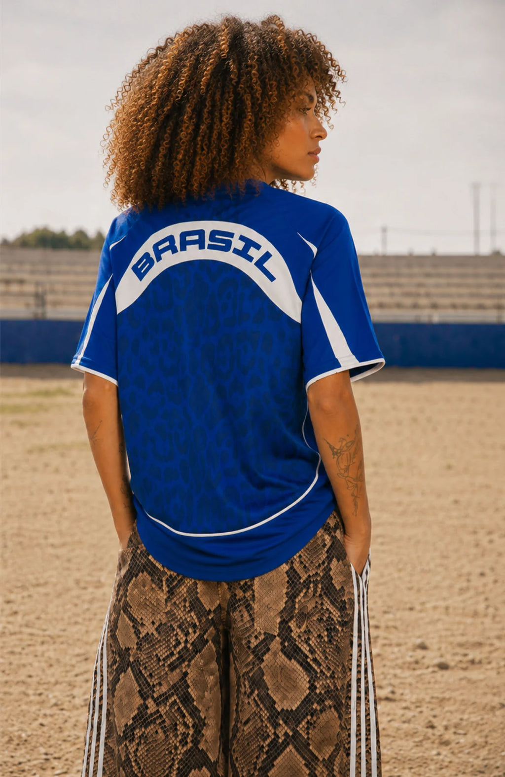 A woman viewed from behind wearing a Varldens royal blue athletic jersey with 'BRASIL' printed across the back, paired with brown and beige snakeskin print pants, standing on a sandy field.