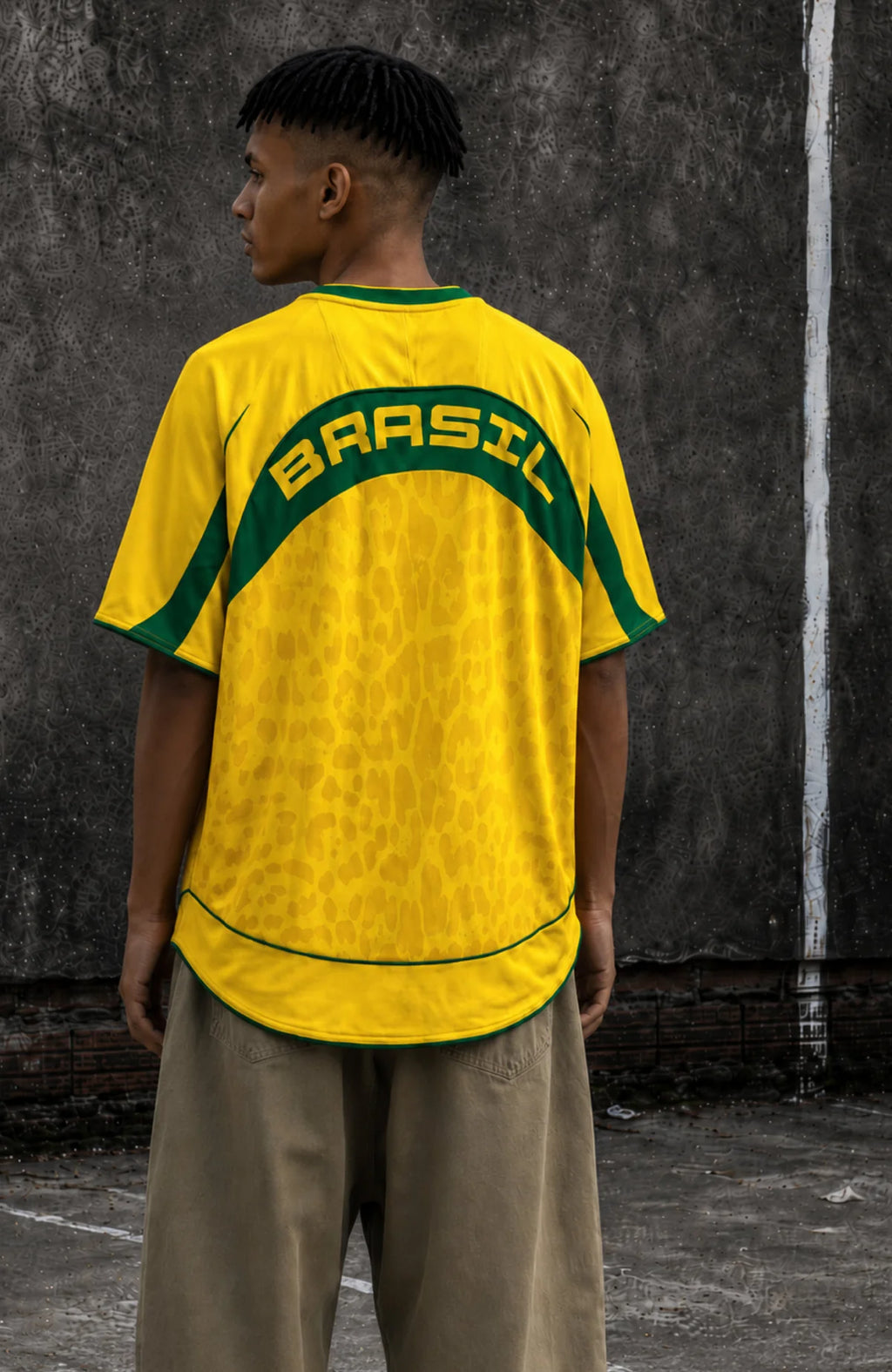 A man wearing a Varldens yellow and green Brazil soccer jersey with 'BRASIL' printed across the back, paired with beige pants, standing against a textured concrete wall.