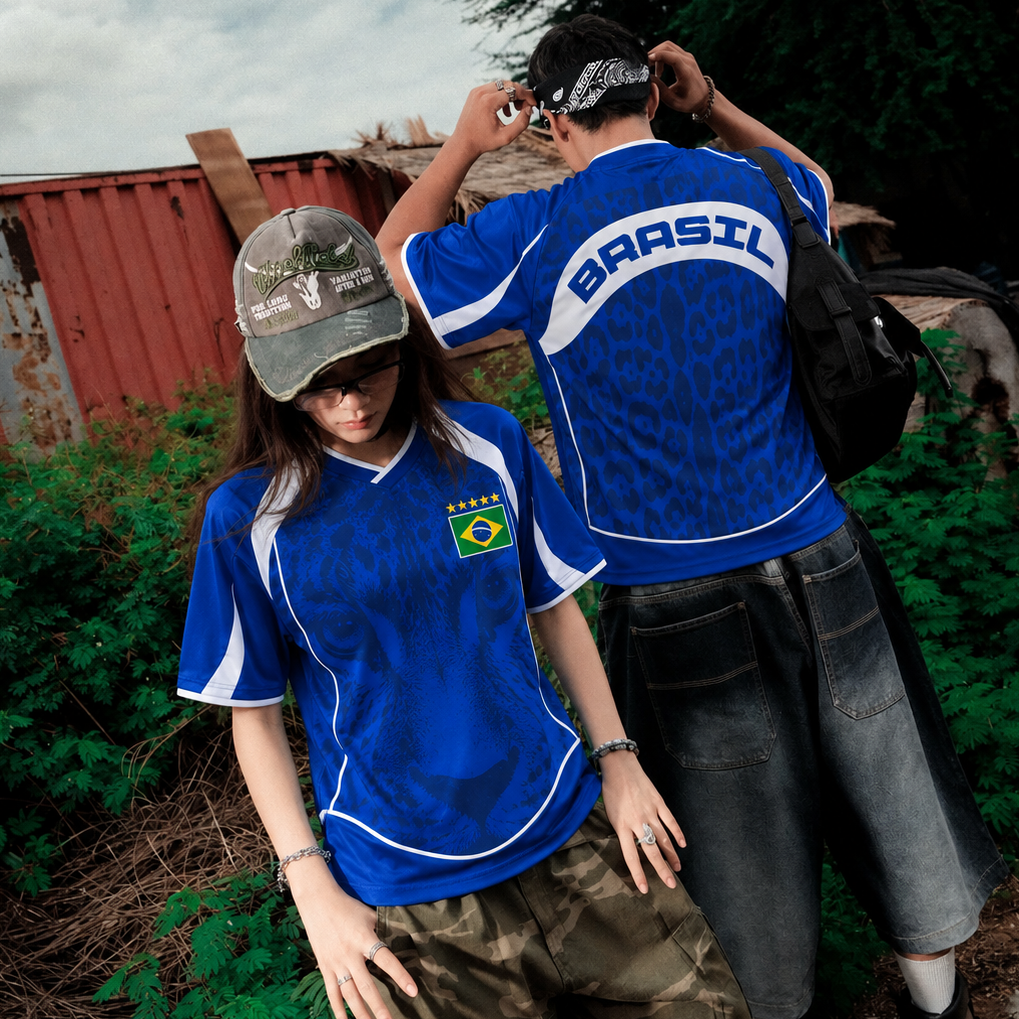 Two models wearing blue Brazil soccer jerseys in an outdoor rustic setting. The model in the foreground wears a gray cap and camouflage cargo shorts, while the model in the background wears a black bandana and dark denim shorts. Varldens