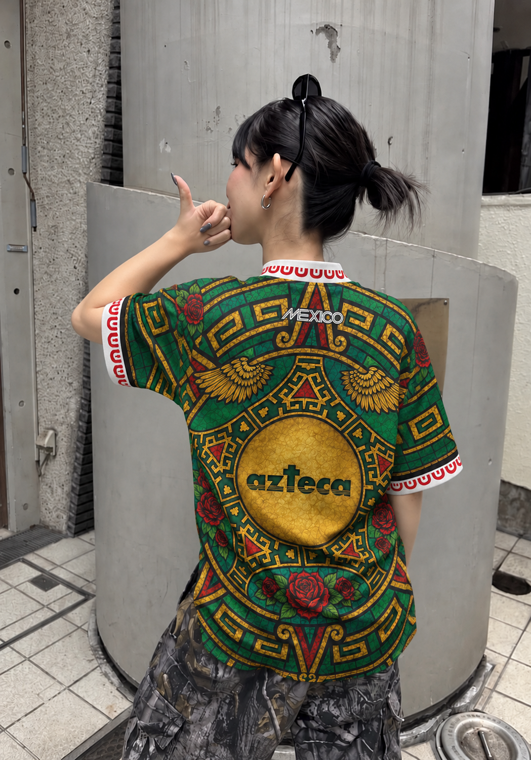 A person shown from behind wearing a colorful Azteca Mexica graphic t-shirt with a green, gold, and red design, standing outdoors next to a concrete structure. Varldens