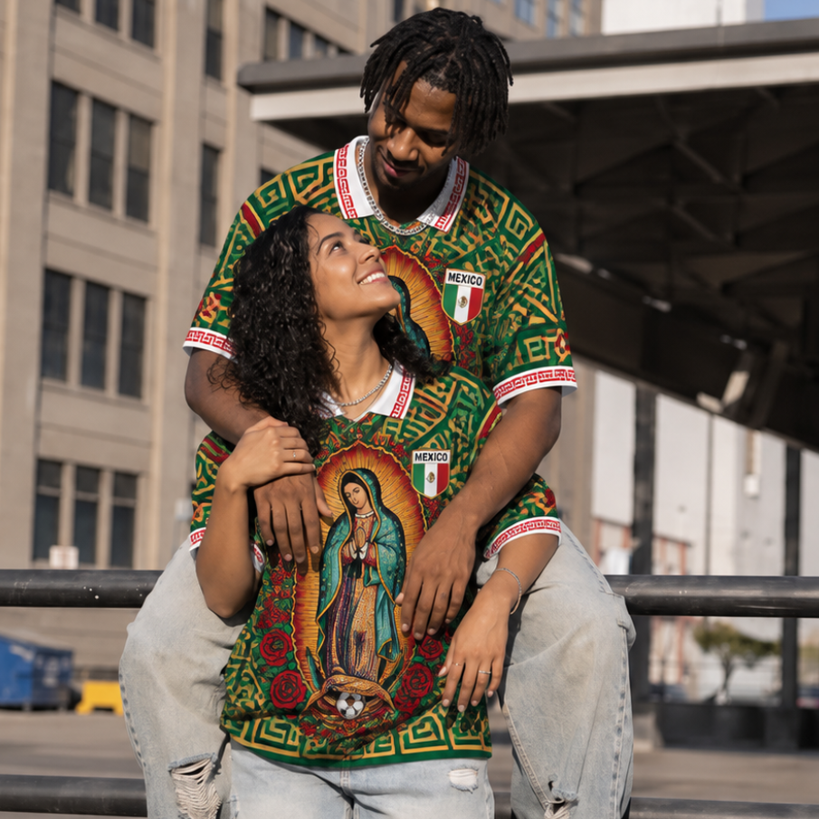 A couple wearing matching green Mexican-themed graphic jerseys with distressed light-wash jeans of Varldens, standing outdoors in an urban setting. The man has his legs crossed over the woman, and both shirts feature a colorful religious illustration of Our Lady of Guadalupe, Mexican flag details, and geometric patterns.